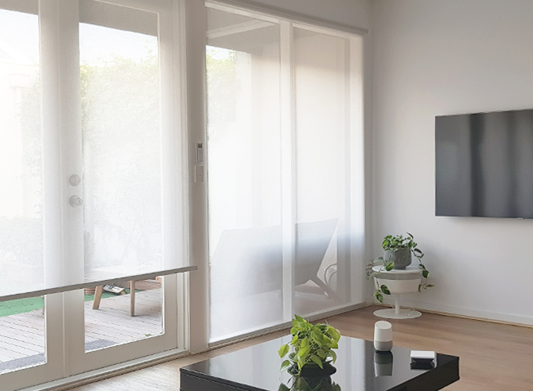 A modern living room featuring automated white sunscreen blinds on the window and french doors, operated by the Google HomePod and wifi hub on the coffee table.