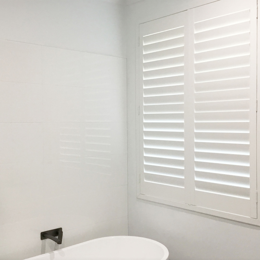 Modern white bathroom with freestanding bath, black taps and a white plantation shutter on the window above the bath