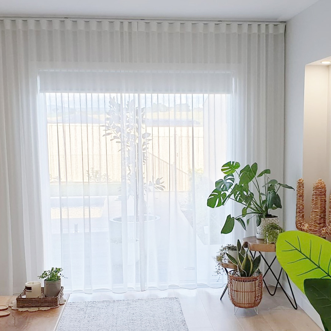 Coastal style living room with white sheer curtains and blockout roller blinds behind, looking out to landscaped backyard.