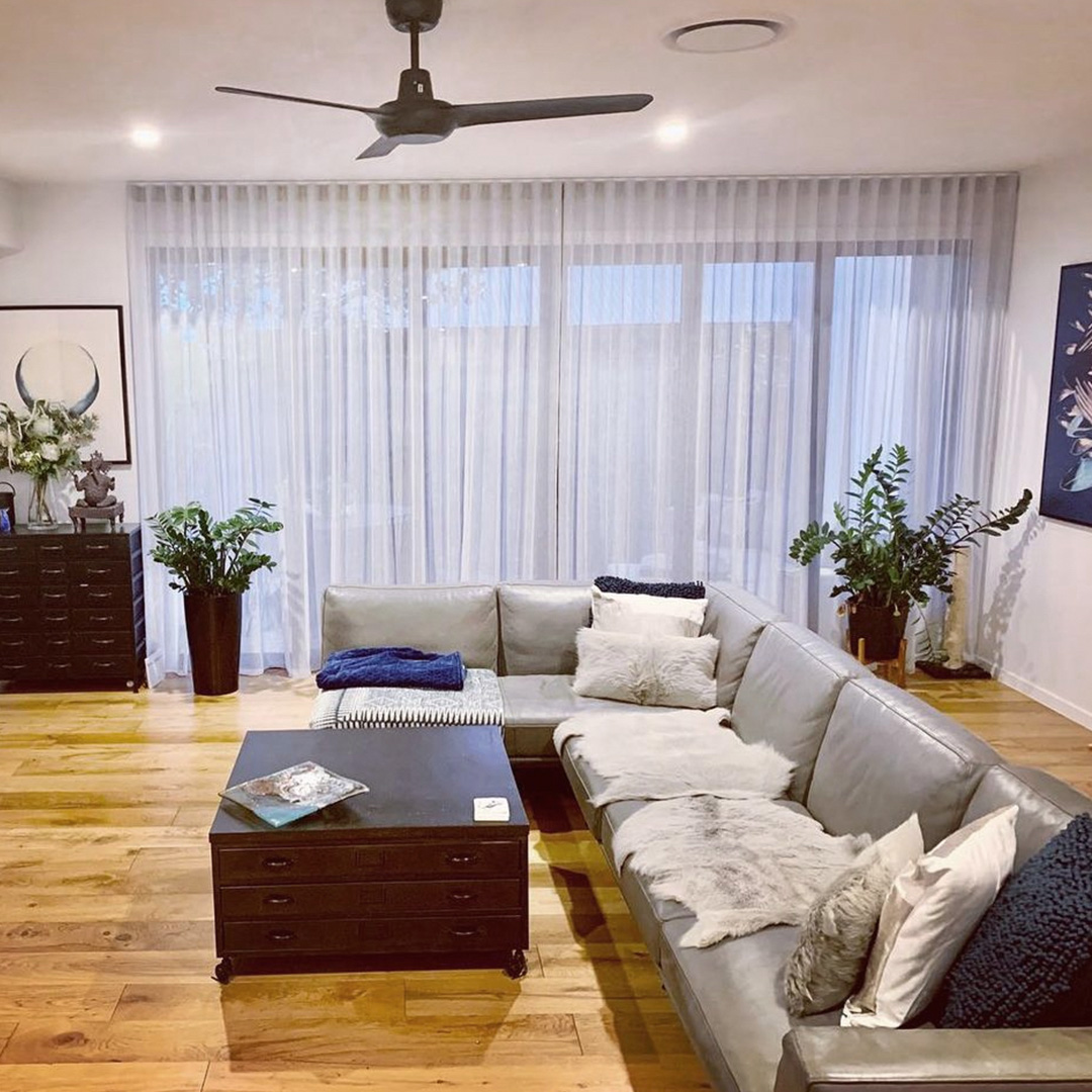 Casual living room with large sofa and light grey sheer curtains on the windows at rear.