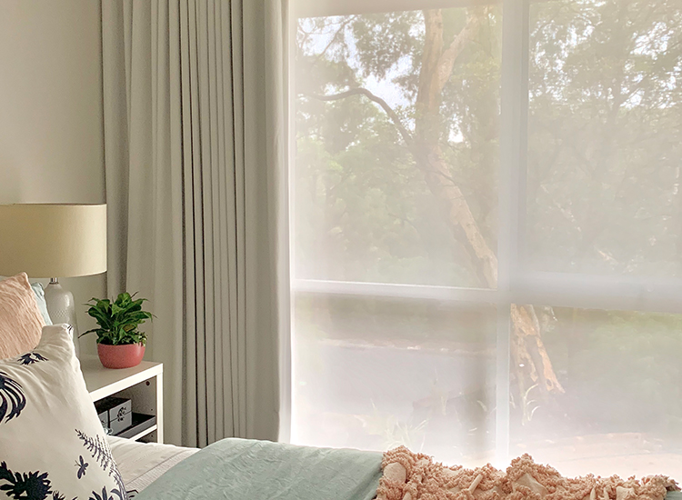 Master bedroom looking across the bed to the white blockout curtain and white sunscreen blind on the window overlooking a garden. 